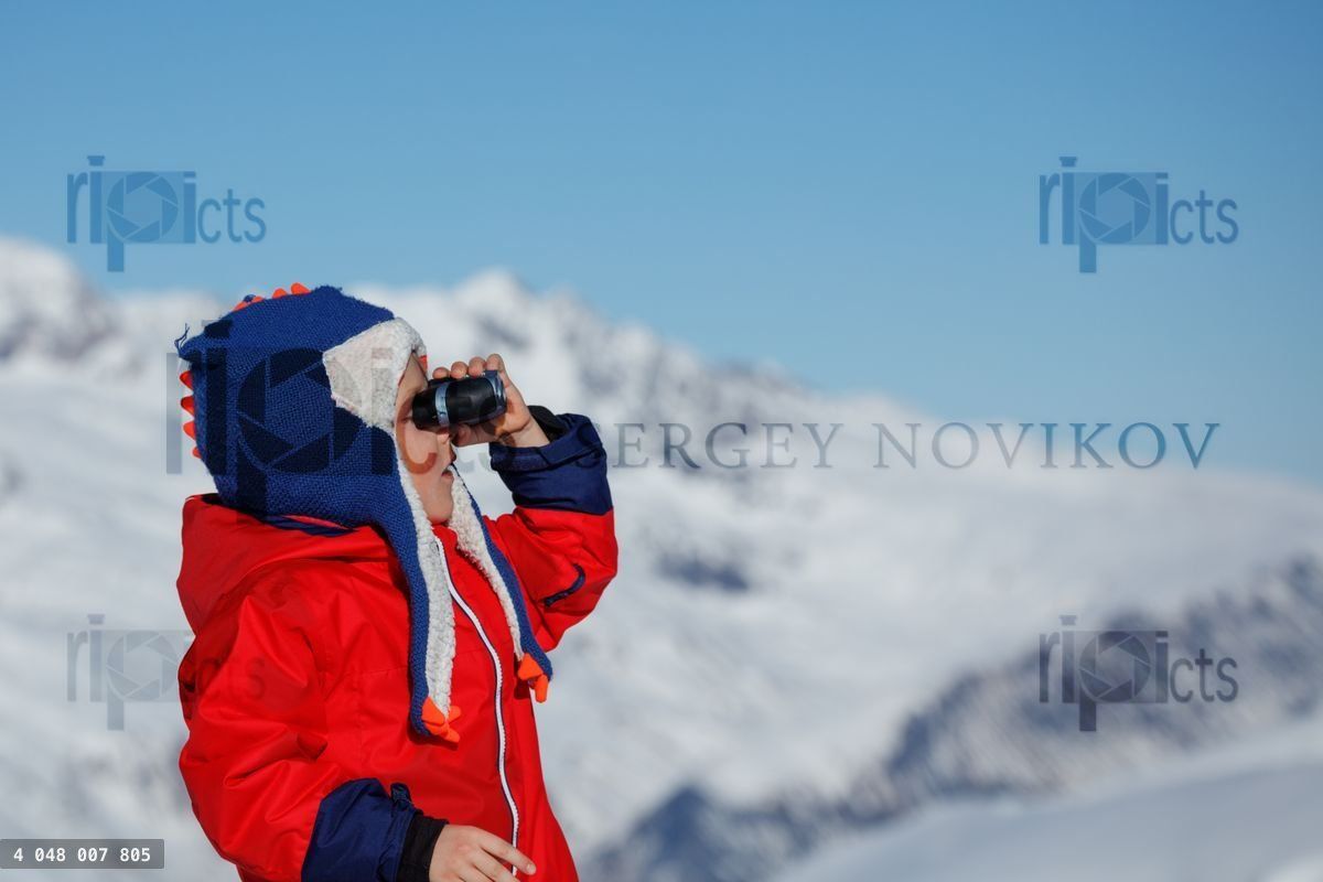 Young kid boy in blue funny hat views snowy peaks at binocular