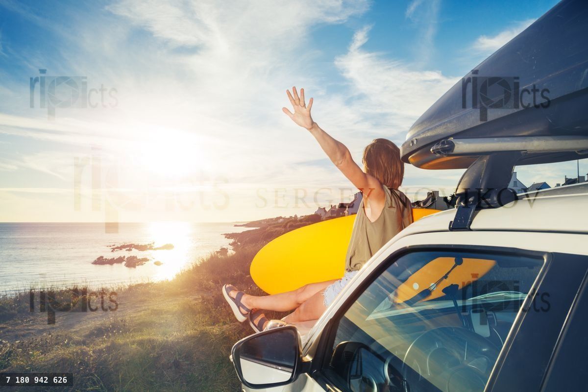 Woman holding surfboard sit on car hood look over sunset