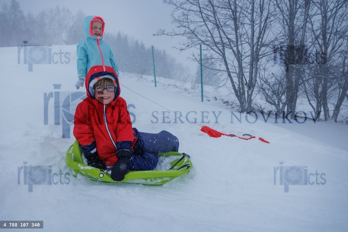 Child and friend sit in green sled smiling on gentle snowy slope
