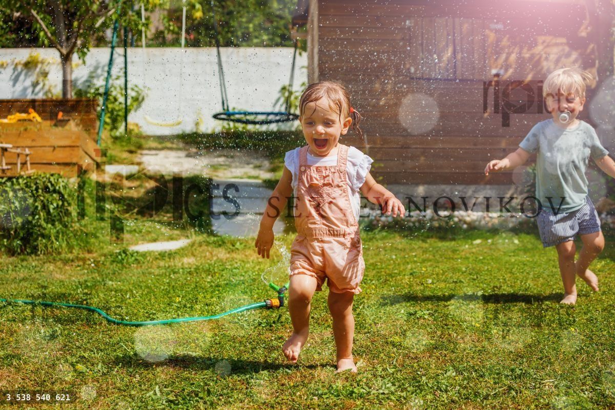 Two happy little children boy, girl run around water sprinkler