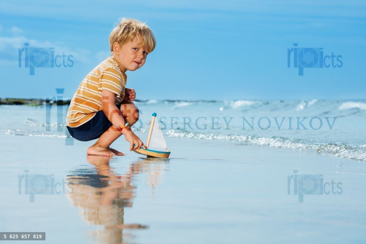 Handsome little blond boy put toy boat in the sea waves water