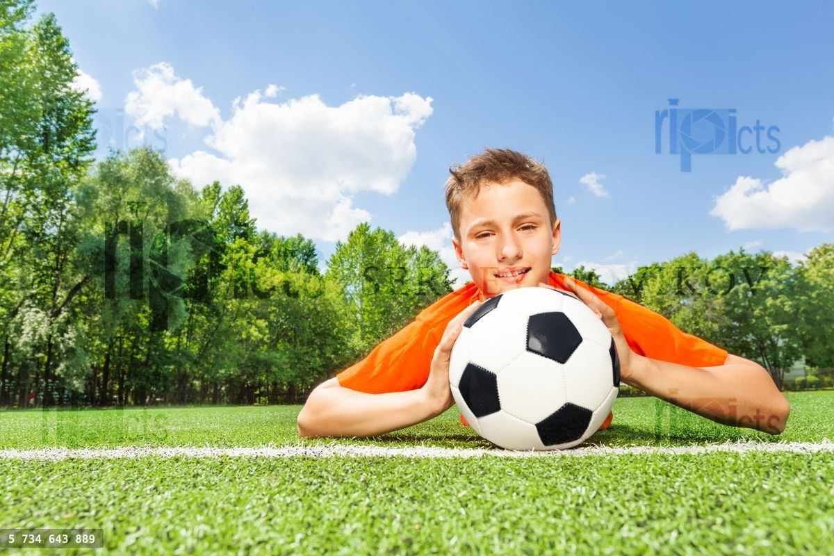 Happy boy holding football lays on green grass