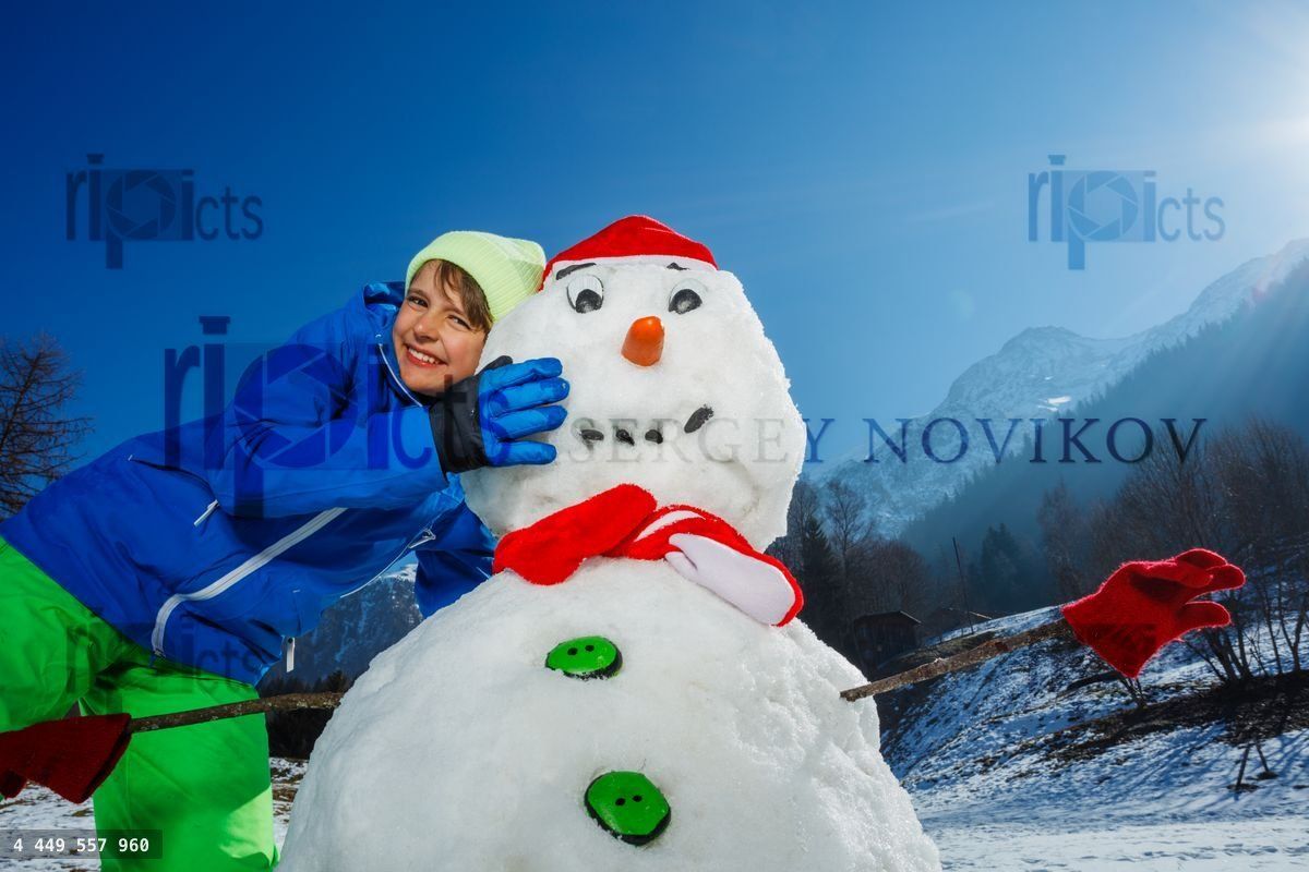 Young boy hugs snowman in winter landscape with mountains