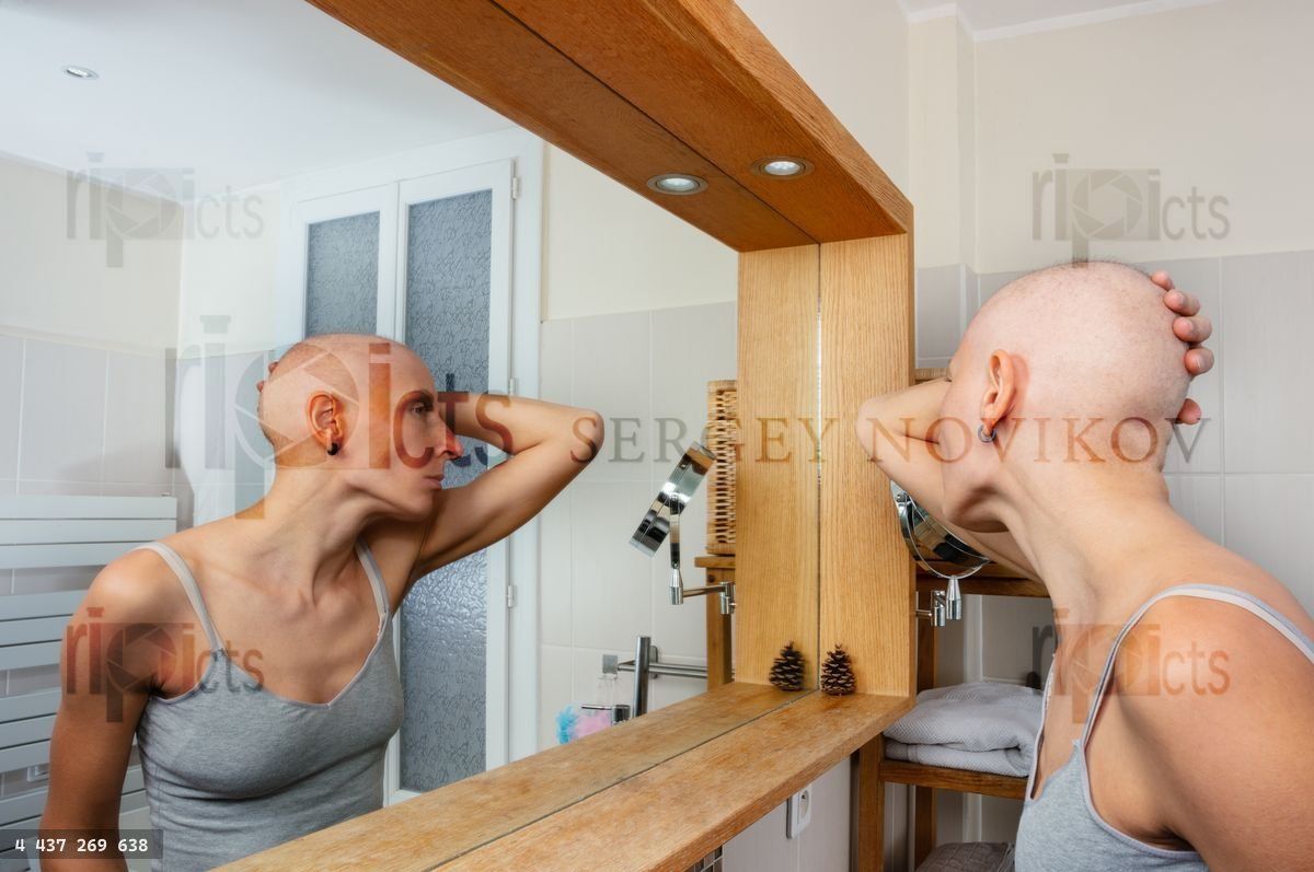 Woman with no hair contemplates her reflection in a mirror