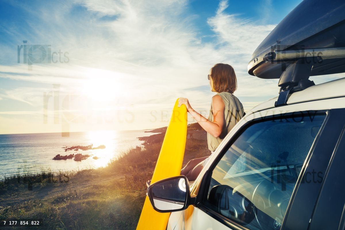 Young woman with surfboard on car hood look at sunset from back
