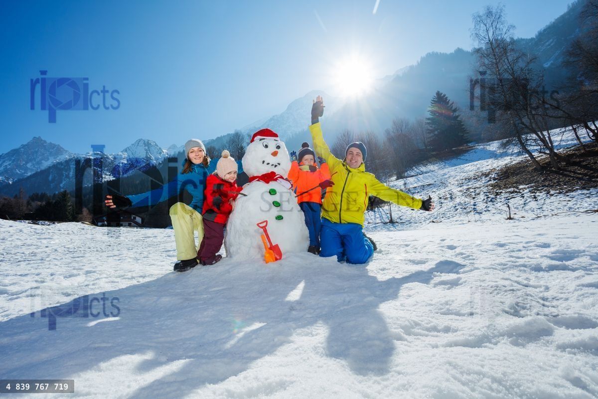 Family with kids in mountains hug snowman wearing Santa hat