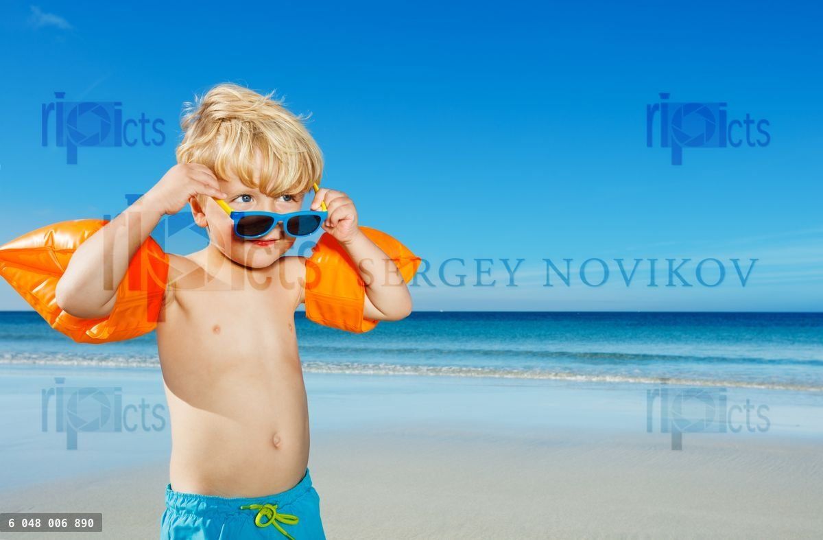 Boy in inflatable shoulder straps holding sunglasses on beach