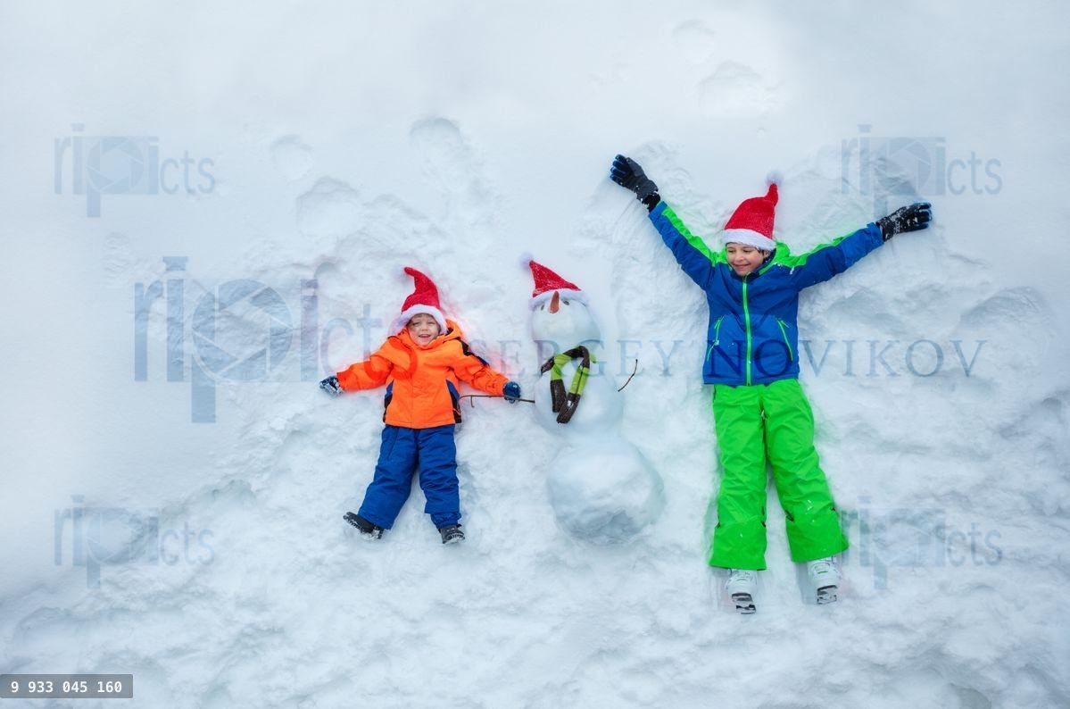 Boys lay in snow together with snowman wear Santa hats top view