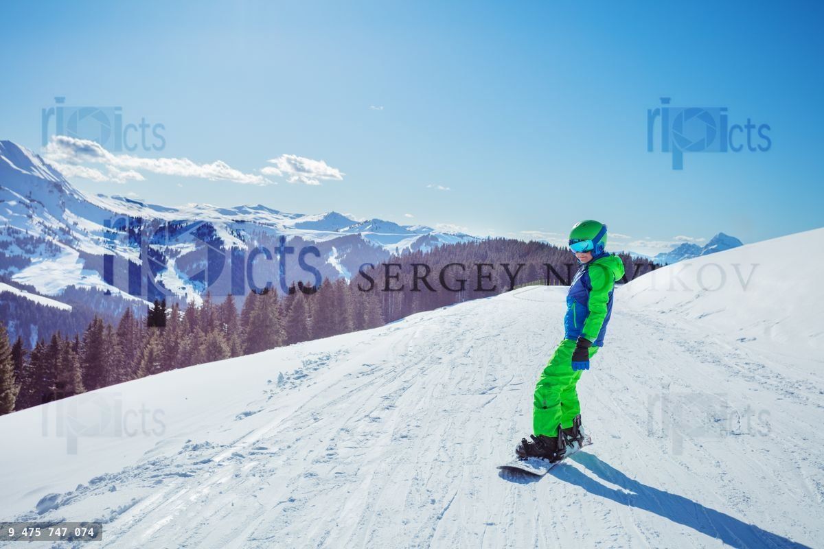 Boy in ski outfit stand on snowboard, sunny view over mountains