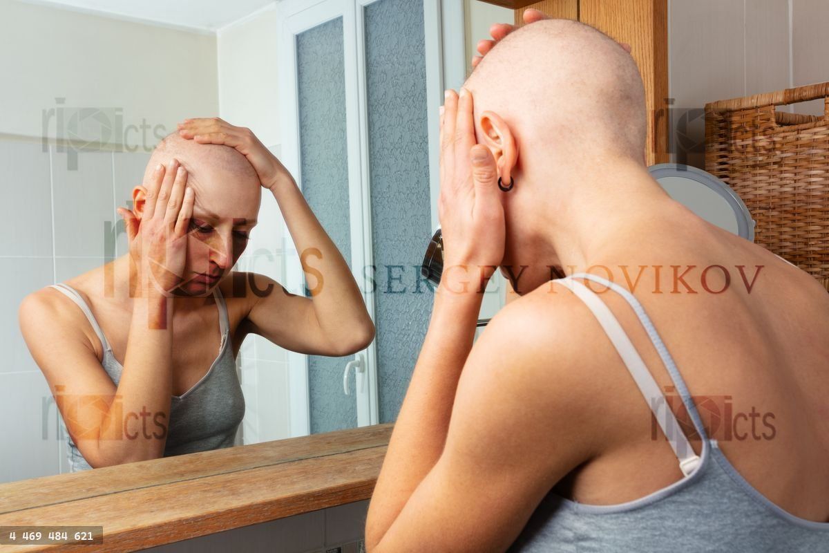 Woman touches shaved head in mirror reflection tired being ill