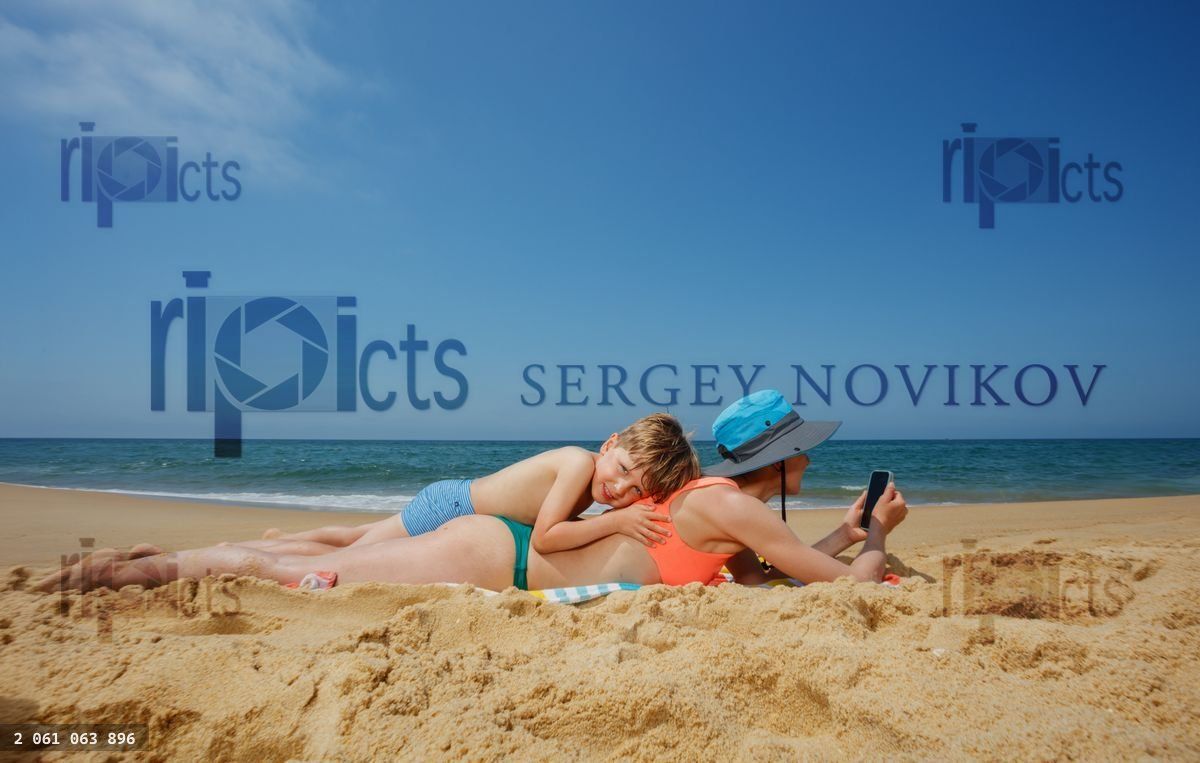 Woman lies on beach and using phone with child resting upon her