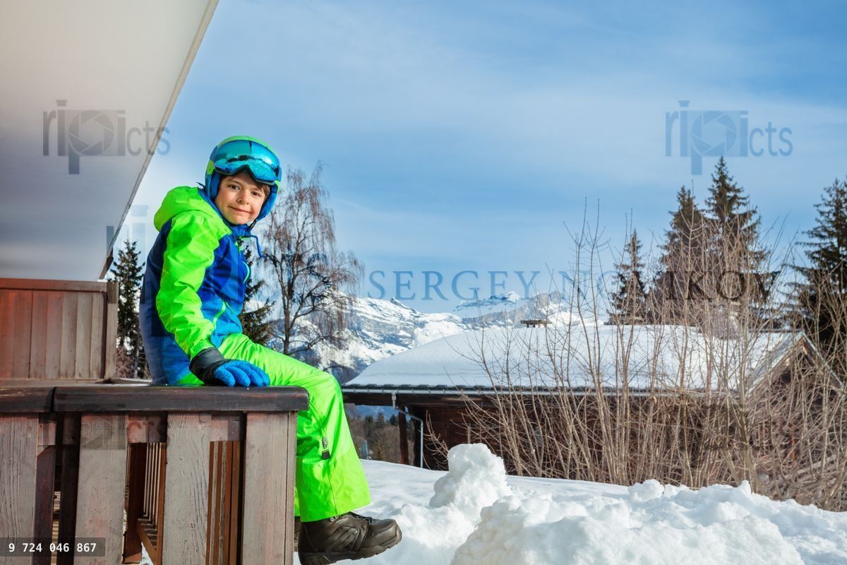 Young child in ski outfit sit on balcony rail at winter house