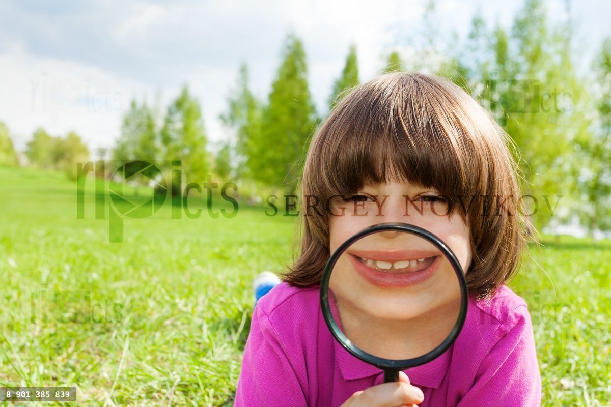 Funny boy smiles through magnifier, lays on grass