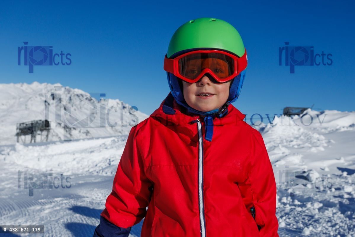 Boy in red jacket and green helmet stands against snowy peaks