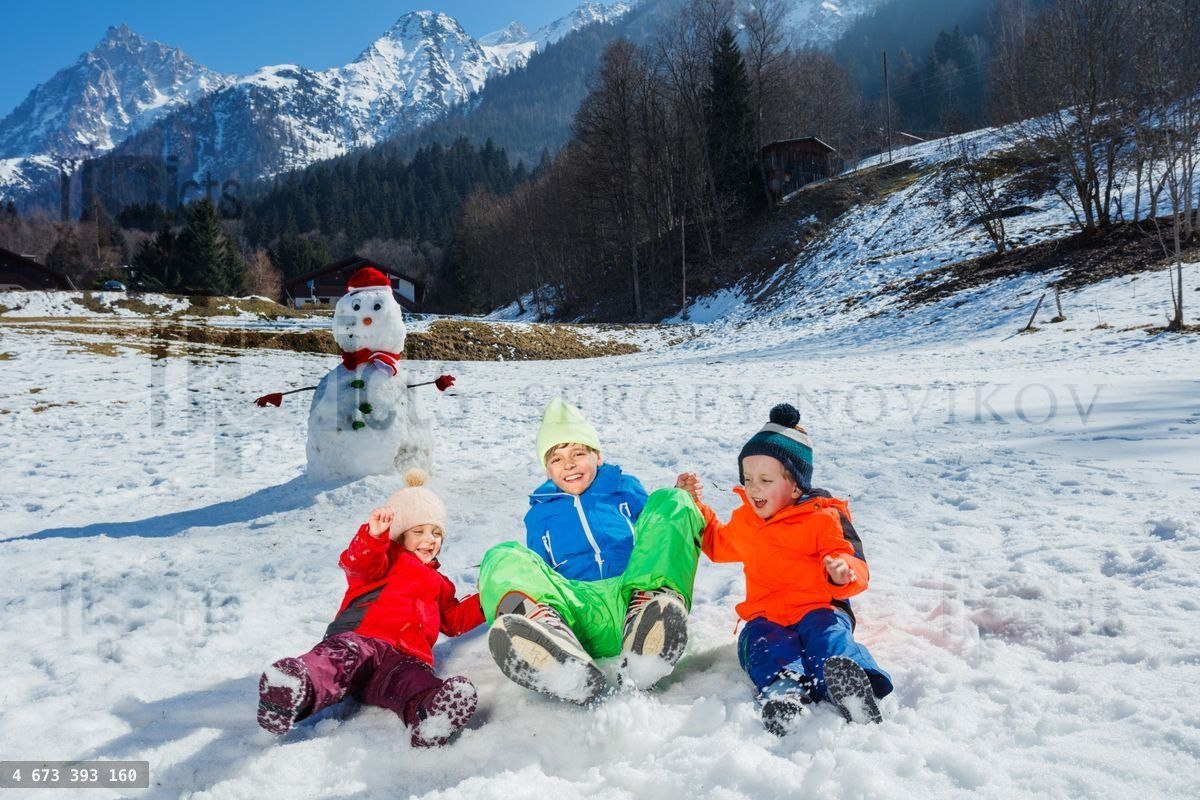 Three children jump together in snow near snowman over mountains
