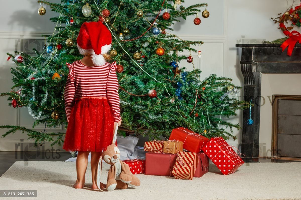 Girl in Santa hat admire Christmas tree and present by fireplace