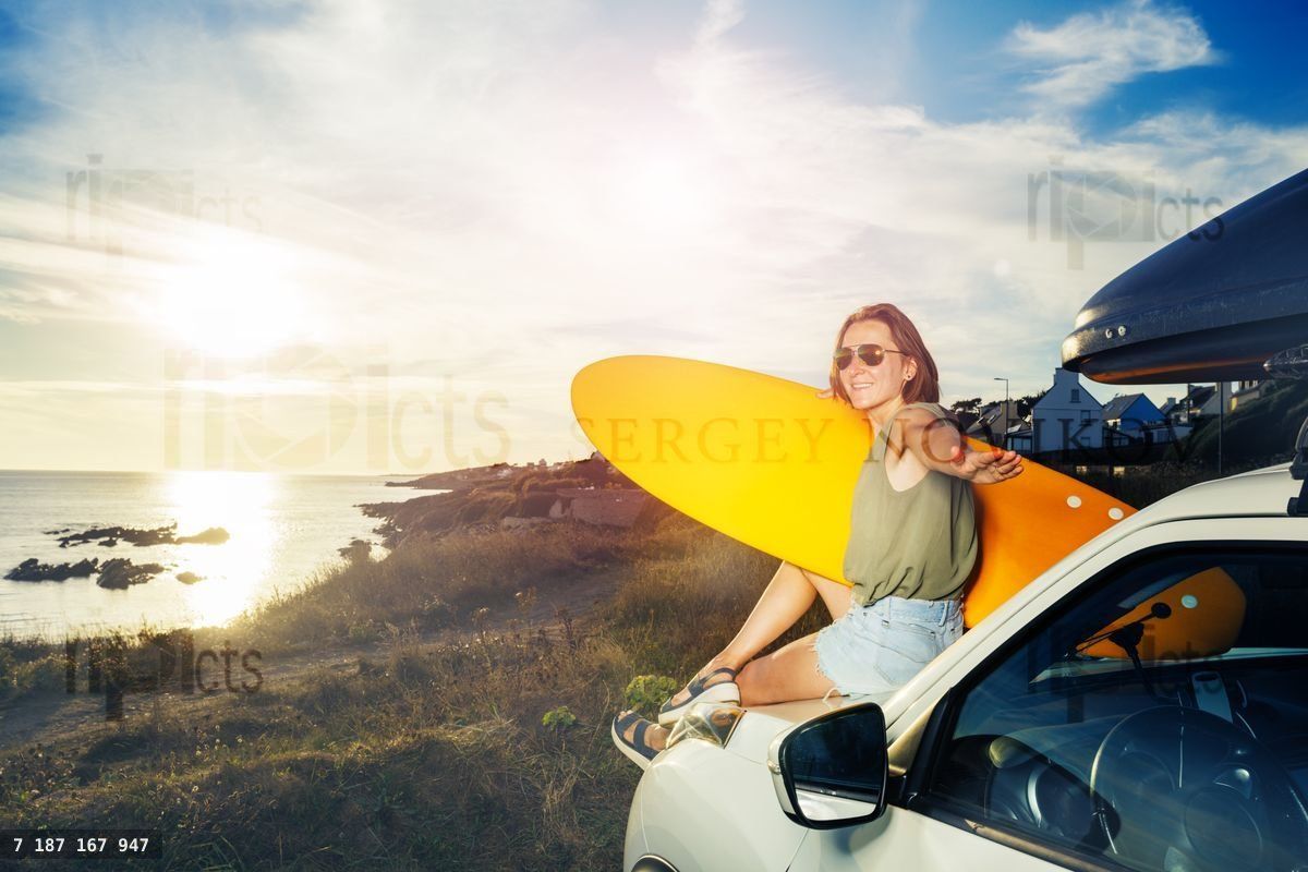 Woman with surfboard sit on car hood look over sunset turn back