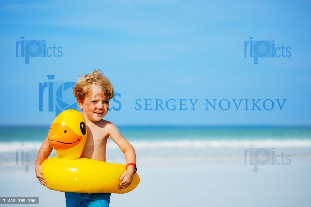Blond boy with inflatable yellow duck buoy stand on sand beach