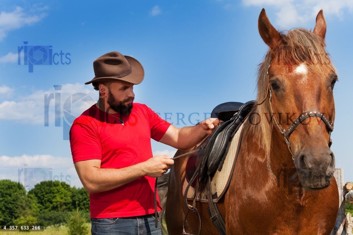 Young man in cowboy hat saddling his brown horse