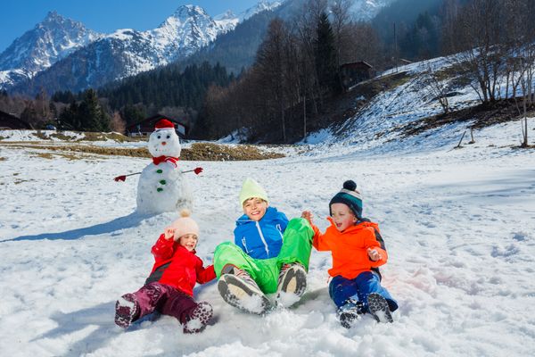 Three children jump together in snow near snowman over mountains